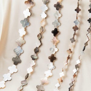 May include: Several strands of decorative clover-shaped beads in various colors, including white, gray, and brown, are displayed against a soft, off-white background. The beads are strung together, creating a flowing, elegant design.