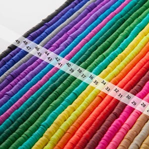 May include: Rows of colorful, flat, disc-shaped beads in a rainbow of hues, including black, purple, blue, green, yellow, orange, and pink. A white ruler with numbers 25-49 is diagonally across the beads. These beads are ideal for jewelry making and crafts.