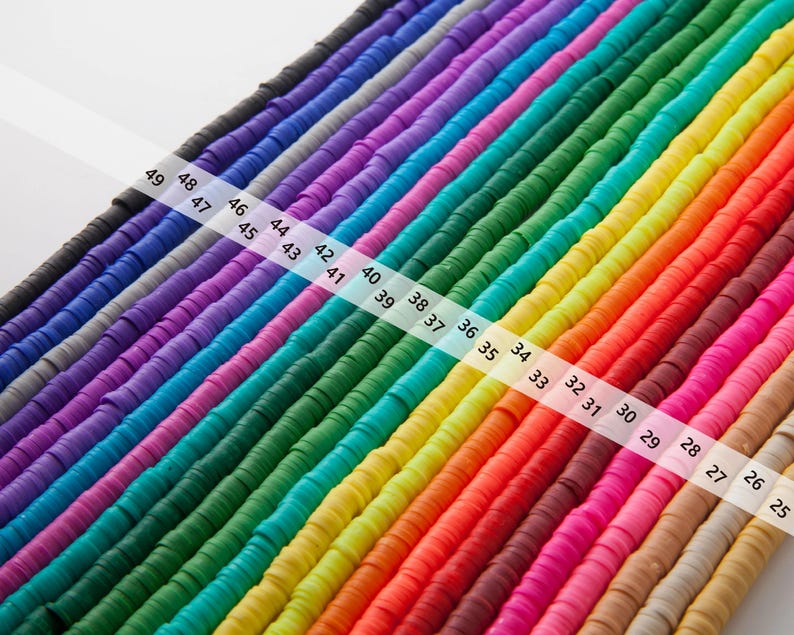 May include: Rows of colorful, flat, disc-shaped beads in a rainbow of hues, including black, purple, blue, green, yellow, orange, and pink. A white ruler with numbers 25-49 is diagonally across the beads. These beads are ideal for jewelry making and crafts.