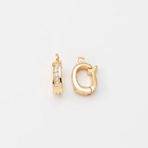 Pode incluir: Duas fechos de joias dourados. Um é um fecho retangular com uma fileira de pedras claras de corte quadrado. O outro é um fecho arredondado com um pequeno laço e um design curvo. Ambos os fechos estão em um fundo branco.