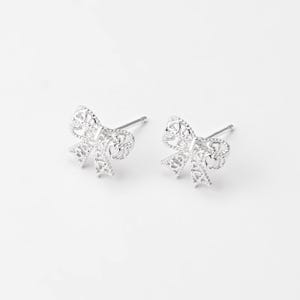 May include: A pair of silver-colored bow-shaped stud earrings. The earrings have a delicate, openwork design with intricate details. The bows are symmetrical and have a polished finish, set against a white background.