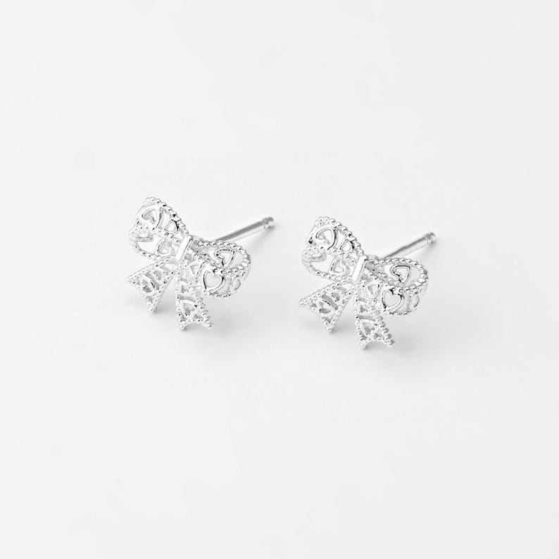 May include: A pair of silver-colored bow-shaped stud earrings. The earrings have a delicate, openwork design with intricate details. The bows are symmetrical and have a polished finish, set against a white background.