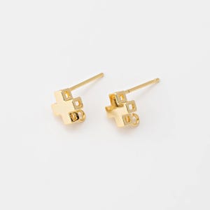 May include: A pair of gold-colored stud earrings. Each earring features a cross-shaped design with square cutouts and a small loop at the bottom. The earrings are set against a plain white background.