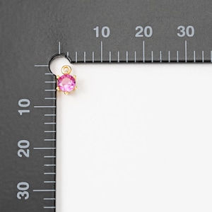 May include: A small pink gemstone charm with a gold setting. The charm is about 0.5 inches in diameter and is shown against a white background with a ruler for scale.