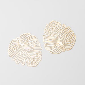May include: Two gold-toned metal charms shaped like monstera leaves. Each charm features a delicate, openwork filigree design that mimics the veins and characteristic split-leaf pattern of a tropical plant, with a small loop at the top.