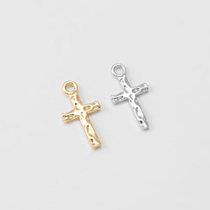 Puede incluir: Dos peque&ntilde;os colgantes de cruz, uno dorado y otro plateado, se muestran sobre un fondo blanco. Cada cruz tiene una superficie texturizada y un lazo en la parte superior para sujetar a una cadena u otro componente de joyer&iacute;a.