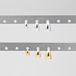 May include: A selection of teardrop-shaped earrings in silver and gold. The earrings are displayed on a grey metal rack. The earrings are of varying sizes, with silver earrings at the top and gold earrings below.