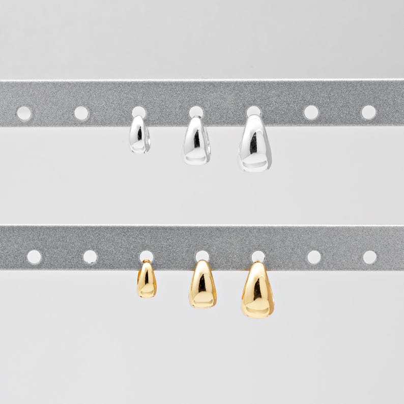 May include: A selection of teardrop-shaped earrings in silver and gold. The earrings are displayed on a grey metal rack. The earrings are of varying sizes, with silver earrings at the top and gold earrings below.