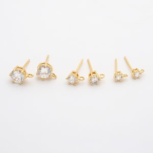 Op de afbeelding: Verschillende paren goudkleurige oorstekers met heldere, ronde, facetgeslepen stenen. De oorbellen variëren in grootte, met een pin en een kleine lus voor het bevestigen van extra elementen. Gepresenteerd op een witte achtergrond.