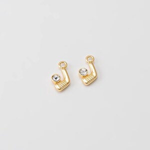 May include: Two gold-colored golf club charms with a clear, round crystal accent. The charms have a loop at the top for attaching to jewelry. The golf club heads have horizontal lines. The background is plain white.