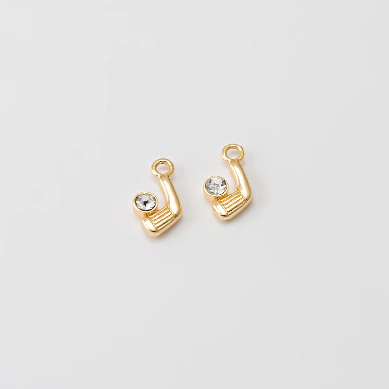 May include: Two gold-colored golf club charms with a clear, round crystal accent. The charms have a loop at the top for attaching to jewelry. The golf club heads have horizontal lines. The background is plain white.