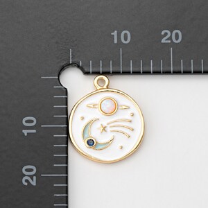 May include: A gold-toned round charm with a white enamel background featuring a celestial design. The charm includes a planet, a crescent moon, a star, and a shooting star. The charm is approximately 0.75 inches in diameter.