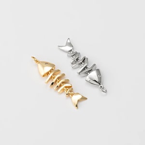 May include: Two fish bone pendants, one gold-toned and one silver-toned, lie on a white surface. The pendants are detailed with articulated segments and a tail fin. The gold pendant has a small, clear gem.