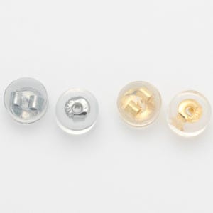 May include: Four earring backs are shown against a white backdrop. Two are silver-coloured metal with clear plastic backings, and two are gold-coloured metal with clear plastic backings. The earring backs are round and designed to secure earrings.