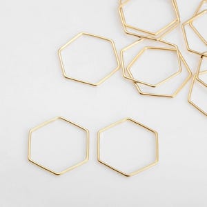 Puede incluir: Una colección de componentes de joyería hexagonales dorados. Las formas de marco abierto están dispersas sobre una superficie blanca, con diferentes tamaños y orientaciones. Los componentes son probablemente para la elaboración de pendientes, colgantes u otras joyas.