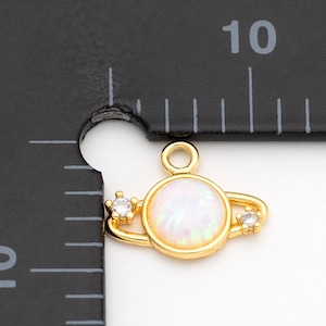 May include: Gold-colored planet charm with a white opal-like center and two small, clear crystals. The charm is approximately 0.4 inches in diameter and has a loop for attaching to jewelry. The charm is shown next to a ruler.
