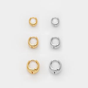 May include: Six small hoop earrings are shown, three gold and three silver, on a white backdrop. The earrings have a rounded, chunky design with a simple clasp. The gold earrings have a polished finish, while the silver ones have a shiny appearance.
