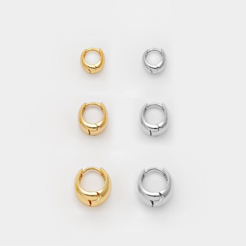 May include: Six small hoop earrings are shown, three gold and three silver, on a white backdrop. The earrings have a rounded, chunky design with a simple clasp. The gold earrings have a polished finish, while the silver ones have a shiny appearance.