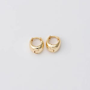 May include: A pair of gold-coloured, chunky hoop earrings. Each earring has a rounded, polished surface and a hinged closure. The earrings are displayed on a white background, highlighting their simple, yet elegant design. These earrings are a classic jewellery accessory.