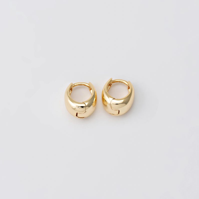 May include: A pair of gold-coloured, chunky hoop earrings. Each earring has a rounded, polished surface and a hinged closure. The earrings are displayed on a white background, highlighting their simple, yet elegant design. These earrings are a classic jewellery accessory.