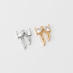 May include: Two bow-shaped charms, one silver-toned and one gold-toned, each adorned with clear, sparkling gemstones. The bows have a central knot and flowing ribbon-like ends, with the silver charm featuring clear stones along the ribbon.