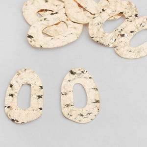 May include: Gold-colored, hammered metal earrings with an oval shape and a central cutout. The earrings have a textured surface and a small hole at the top for attachment. Several other similar pieces are scattered around.