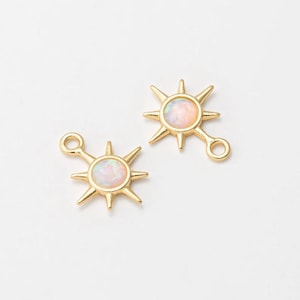 Op de afbeelding: Twee goudkleurige sunburst bedels met een centrale opaalachtige steen. Elke bedel heeft een lus om aan sieraden te bevestigen. Het sunburst-ontwerp heeft puntige stralen die zich uitstrekken vanaf de centrale steen.