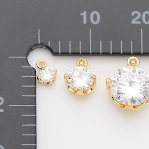 May include: Three gold-colored, round jewelry charms with clear, sparkling center stones. The charms are of varying sizes, with the largest measuring approximately 0.75 inches in diameter. Each charm has a small loop at the top for attaching to a chain or other jewelry.
