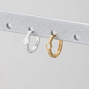 May include: Two hoop earrings, one silver and one gold, each with a small opal stone and encrusted with small clear stones. The earrings are hanging on a silver bar.
