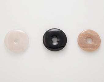 Puede incluir: Tres colgantes de piedra pulida con forma de rosquilla. Uno es de color rosa claro, otro negro y el tercero marrón claro con aspecto veteado. Cada colgante tiene un agujero circular en el centro.