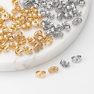 May include: A selection of gold and silver earring backs, presented on a white surface. The earring backs are in various shapes and sizes, with a focus on the butterfly style. The gold backs have a shiny finish, while the silver backs are more matte.