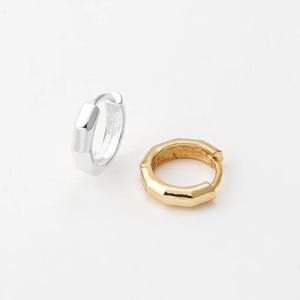 Op de afbeelding: Twee kleine oorringen. Eén is zilverkleurig, de andere goudkleurig. Beide oorbellen hebben een geometrisch, facetgeslepen ontwerp. De oorbellen worden op een witte achtergrond getoond.