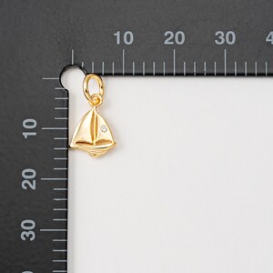 May include: A gold sailboat charm with a small clear rhinestone in the center. The charm is hanging from a ruler with markings in inches.