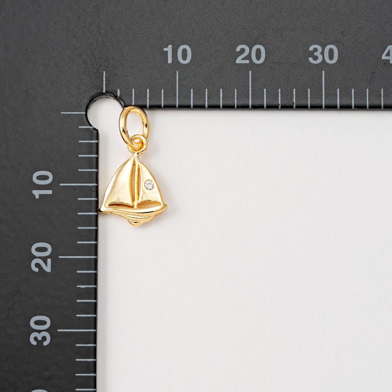 May include: A gold sailboat charm with a small clear rhinestone in the center. The charm is hanging from a ruler with markings in inches.