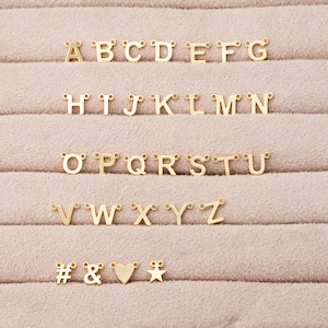 Op de afbeelding: Goudkleurige alfabetletterbedels, inclusief letters A tot en met Z, evenals een nummerteken, een ampersand, een hart en een ster. De bedels zijn gerangschikt in rijen op een lichtbruin, getextureerd oppervlak.