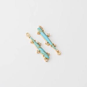 May include: Two gold-toned, elongated pendant charms. Each charm features a turquoise-colored, marbled design with clear crystal accents. The charms are approximately 2 inches long and have a loop at one end.