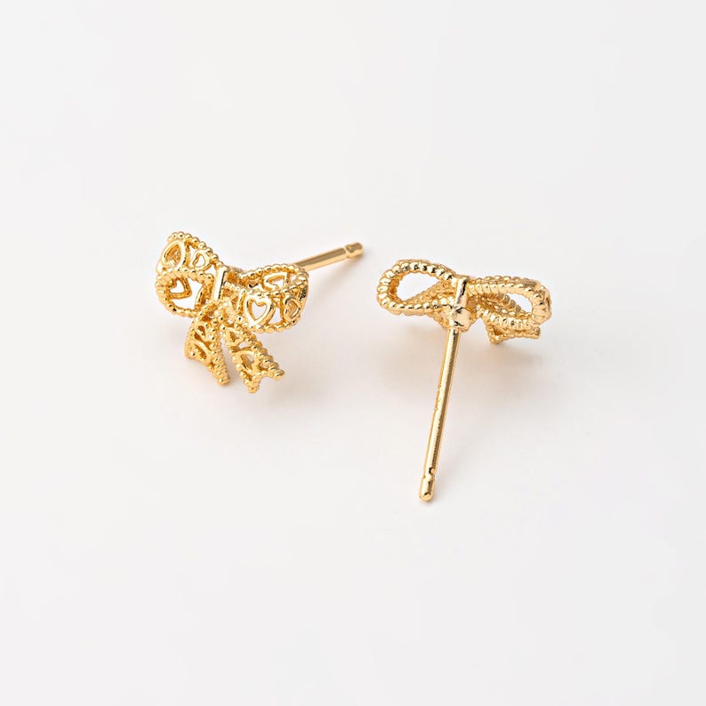 May include: A pair of gold-colored bow-shaped stud earrings. The earrings feature a detailed, openwork design with heart-shaped cutouts. The bows are on gold posts, set against a white background.