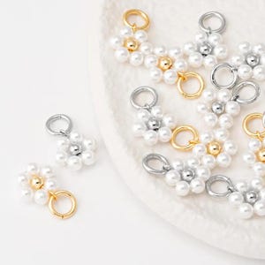 Puede incluir: Primer plano de varios dijes con forma de flor. Cada dije está hecho de perlas de imitación blancas y presenta un anillo y una cuenta central plateados o dorados. Los dijes están dispuestos sobre una superficie blanca ligeramente texturizada.