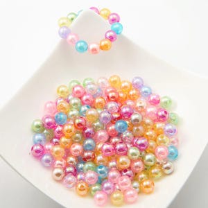 Può includere: Una collezione di perline iridescenti dai colori pastello, tra cui rosa, blu, giallo e verde. Alcune perline sono disposte a forma di braccialetto, mentre altre sono sparse su un piatto quadrato bianco.