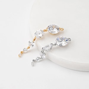 Peut inclure: Deux bijoux pendants élégants. L'un est doré, l'autre argenté. Les deux présentent une série de pierres précieuses claires et étincelantes de différentes formes et tailles. Les pierres précieuses sont disposées dans un design fluide et asymétrique, créant un look sophistiqué.