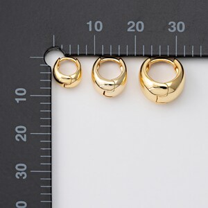 May include: Three gold-coloured hoop earrings in different sizes. The earrings have a shiny, rounded design. The smallest earring is about 1 cm in diameter, the middle one is 1.5 cm, and the largest is 2 cm.