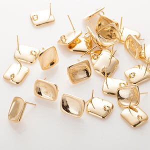 May include: A collection of gold-coloured, square-shaped stud earrings. Each earring has a post for insertion and a slightly rounded face. The earrings are scattered on a white surface, reflecting light.