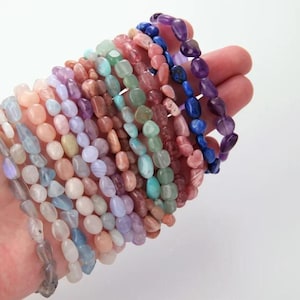 May include: A selection of gemstone bracelets in a variety of colours, including pink, blue, green, and purple. The bracelets are made of polished, irregularly shaped beads. The bracelets are held in a hand against a white background.