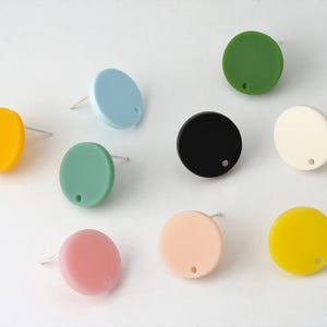 May include: A collection of colorful, round stud earrings in various shades including yellow, blue, green, black, white, pink, and peach. Each earring has a small hole and a silver post. The earrings are arranged on a white surface.