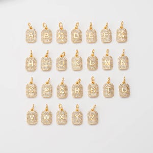Op de afbeelding: Een verzameling goudkleurige rechthoekige hangers, elk versierd met een letter van het alfabet, van A tot Z. De hangers zijn ingelegd met kleine, sprankelende steentjes en hebben een kleine lus aan de bovenkant voor bevestiging.