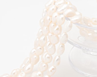 May include: Close-up of creamy white freshwater pearls. The pearls are irregularly shaped and have a lustrous sheen. The pearls are draped over a clear glass container. The background is a clean white.