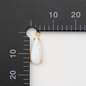 May include: A teardrop-shaped white opal pendant set in a gold-toned metal prong setting. The pendant is positioned against a black measuring tool with white markings, showing a length of approximately 15 millimeters and a width of 7 millimeters.