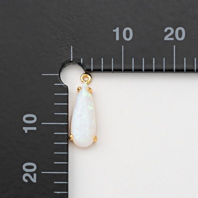 May include: A teardrop-shaped white opal pendant set in a gold-toned metal prong setting. The pendant is positioned against a black measuring tool with white markings, showing a length of approximately 15 millimeters and a width of 7 millimeters.