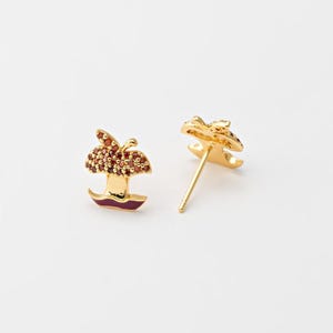 以下が含まれることがあります： 金色のリンゴスタッドピアス。リンゴのデザインは、赤い宝石のクラスター、金の茎、赤いエナメルベースが特徴です。ピアスは白い背景にセットされています。
