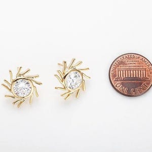 Op de afbeelding: Twee goudkleurige oorstekers met een zonnestraalontwerp, elk met een heldere, ronde edelsteen in het midden. Een Amerikaanse munt ligt naast de oorbellen ter vergelijking. De oorbellen staan op een witte achtergrond.